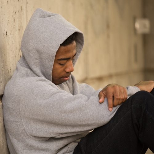 Black male teenage years sits against a concrete wall. He has a grey hoody on and it is covering his head. He has black pants on. His gaze is downwards and he looks sad and depressed.