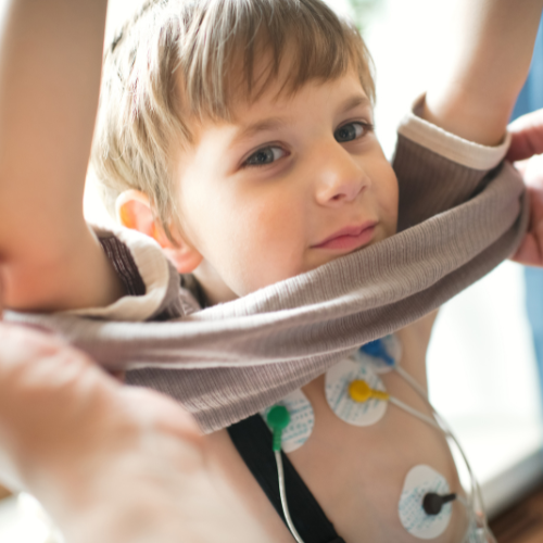 Young boy with short blond hair is putting on a shirt. Atttached to his chest is a holter monitor patches that are tracking his heart rate.